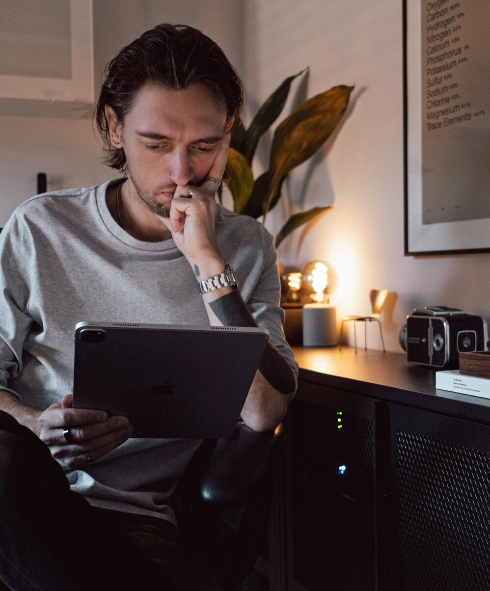 A photograph of a minimal wooden desk in a bright room, featuring a wall-mounted monitor, a tall, potted cactus, and a large, white desktop speaker.